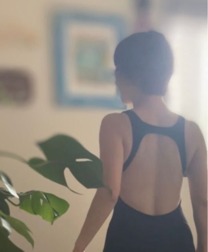 黒いワンピースを着た女性が、観葉植物のそばに立って後ろ姿を見せている。背景には絵画が飾られた壁がある。
