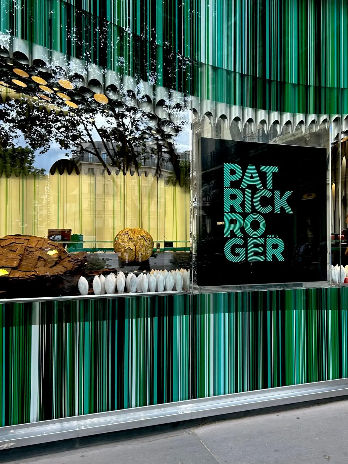 パトリックロジェの緑色の縞模様のガラス窓が特徴の店頭で、店内には人々が集まり、テラスには白いカップと装飾的な陶器が並んでいる。