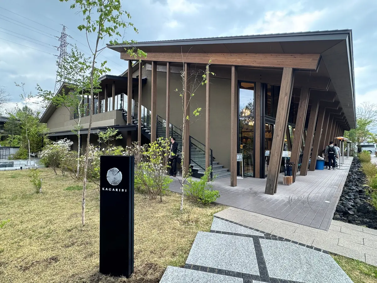 木造のモダンな建物と芝生、木々のある屋外空間。建物の前には「KAKARIBI」の看板が立つ。