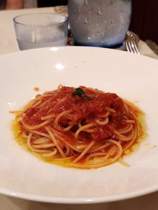 白い皿に盛り付けられたスパゲッティ。トマトソースがたっぷりかかり、緑の葉が添えられている。背景にはグラスとフォークが置かれている。