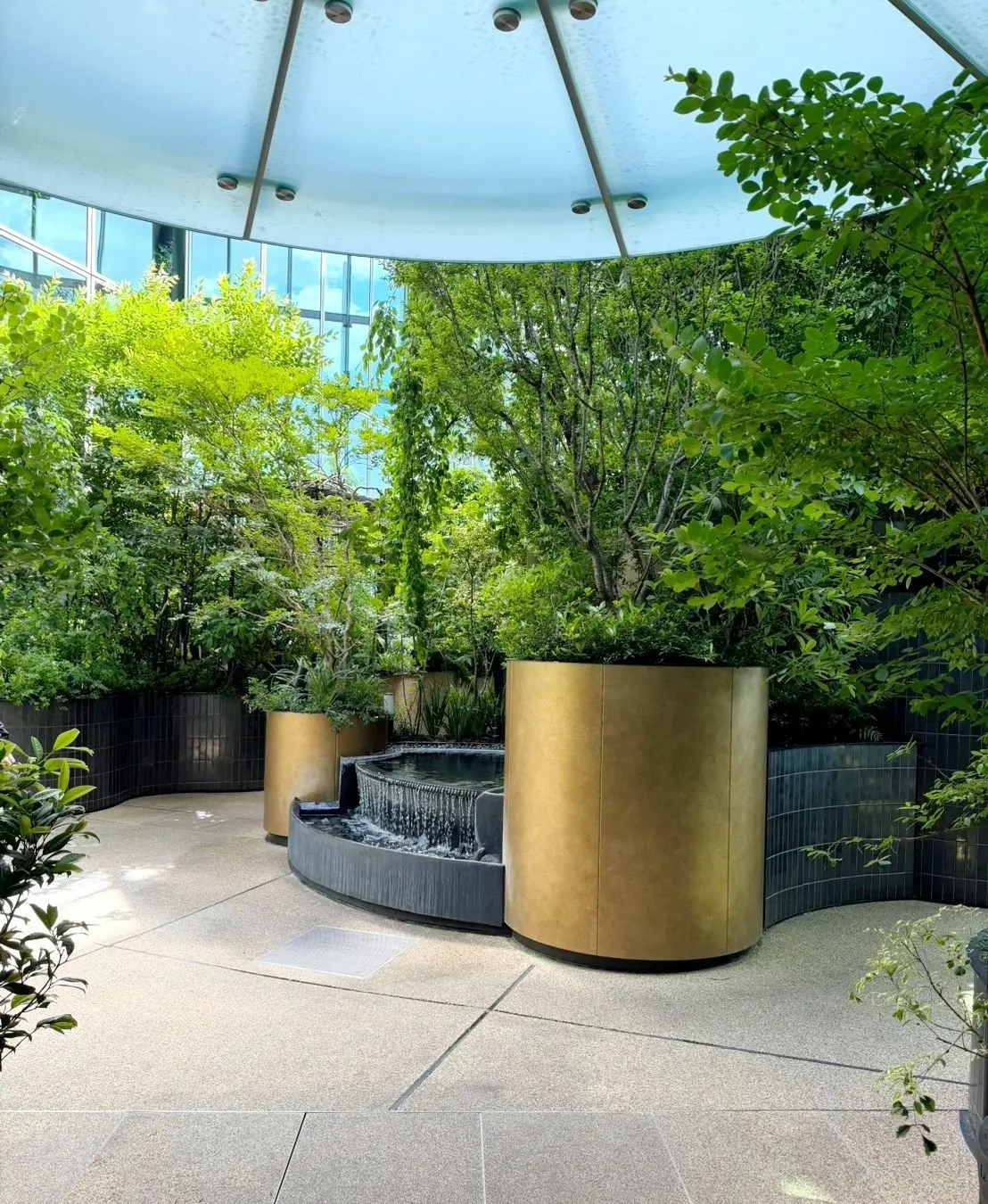 屋内庭園。中央に水が流れる金色の水盤があり、その周りに大型の植木鉢に入った緑豊かな植物が配置されている。半透明の屋根が設置され、自然光が差し込んでいる。
