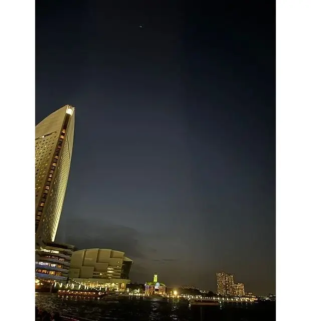夜の横浜港、高層ビルと複合施設がライトアップされ、水面に反射している。空には月が小さく浮かんでいる。