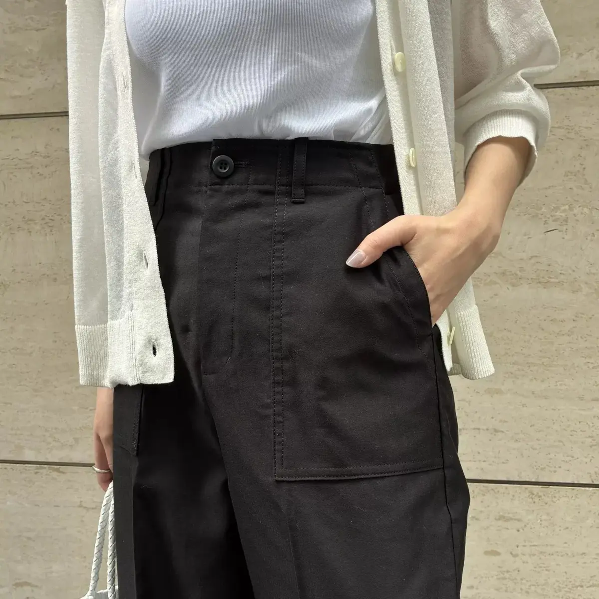 白のTシャツにオフホワイトのカーディガン、黒いカーゴパンツを着用した女性。右手をポケットに入れ、正面を向いている。背景はグレーのコンクリート壁。