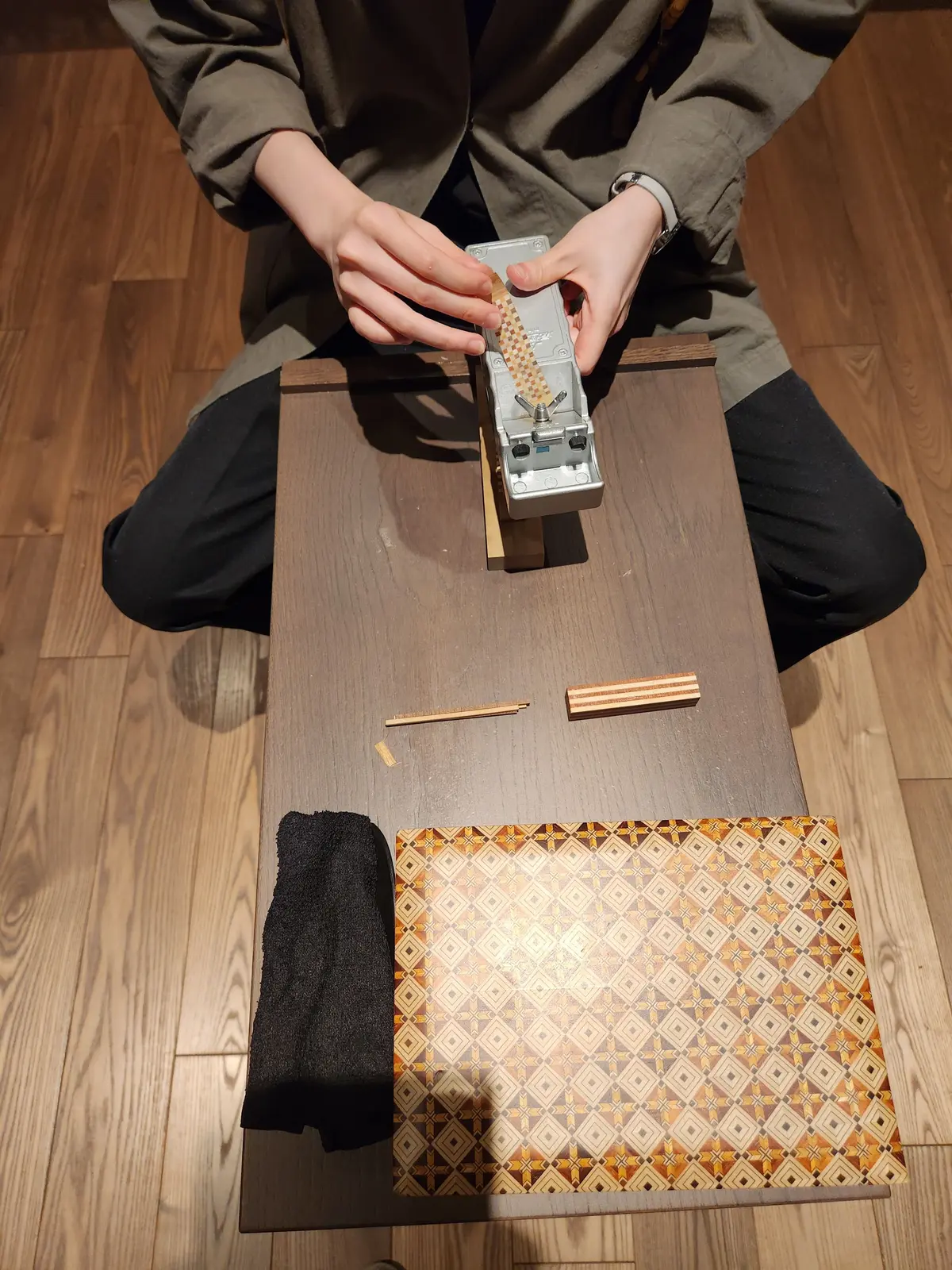 木製のテーブルに座る人物が、ラタン製の編み込みパーツを加工する機械を操作している。テーブルの上には、編み込みパーツ、黒い布、模様の入った紙が置かれている。