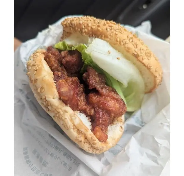 鶏肉の唐揚げが挟まれた、白ゴマ付きのパンのサンドイッチ。レタスが添えられている。紙に包まれており、背景には黒いものが写っている。