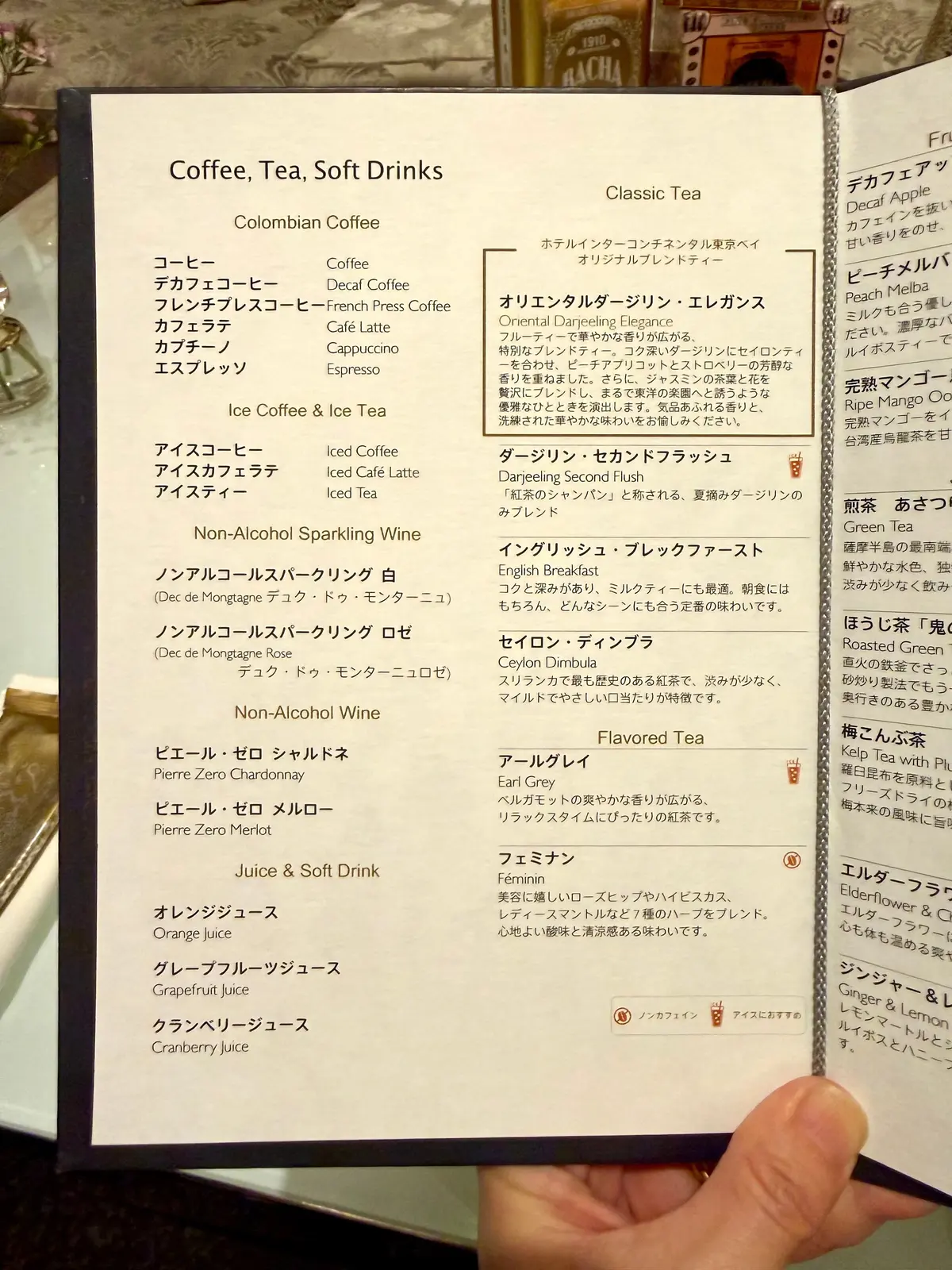 アムステルダムのカフェメニュー。左上に「HEMA」のロゴ。コーヒー、ソフトドリンク、紅茶、フレーバーティー、その他ドリンクの品名と説明が日本語で記載。右ページには、フルーツやハーブを使ったフレーバーティーの品名が記載。背景は茶色と白のストライプ模様。