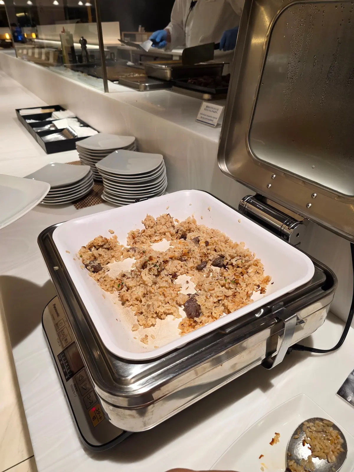 温かいご飯の上に、紫蘇や梅肉と思われる具材が乗った、中華風の炊き込みご飯。ステンレス製の保温容器に入っており、その横には皿が積み重ねられている。