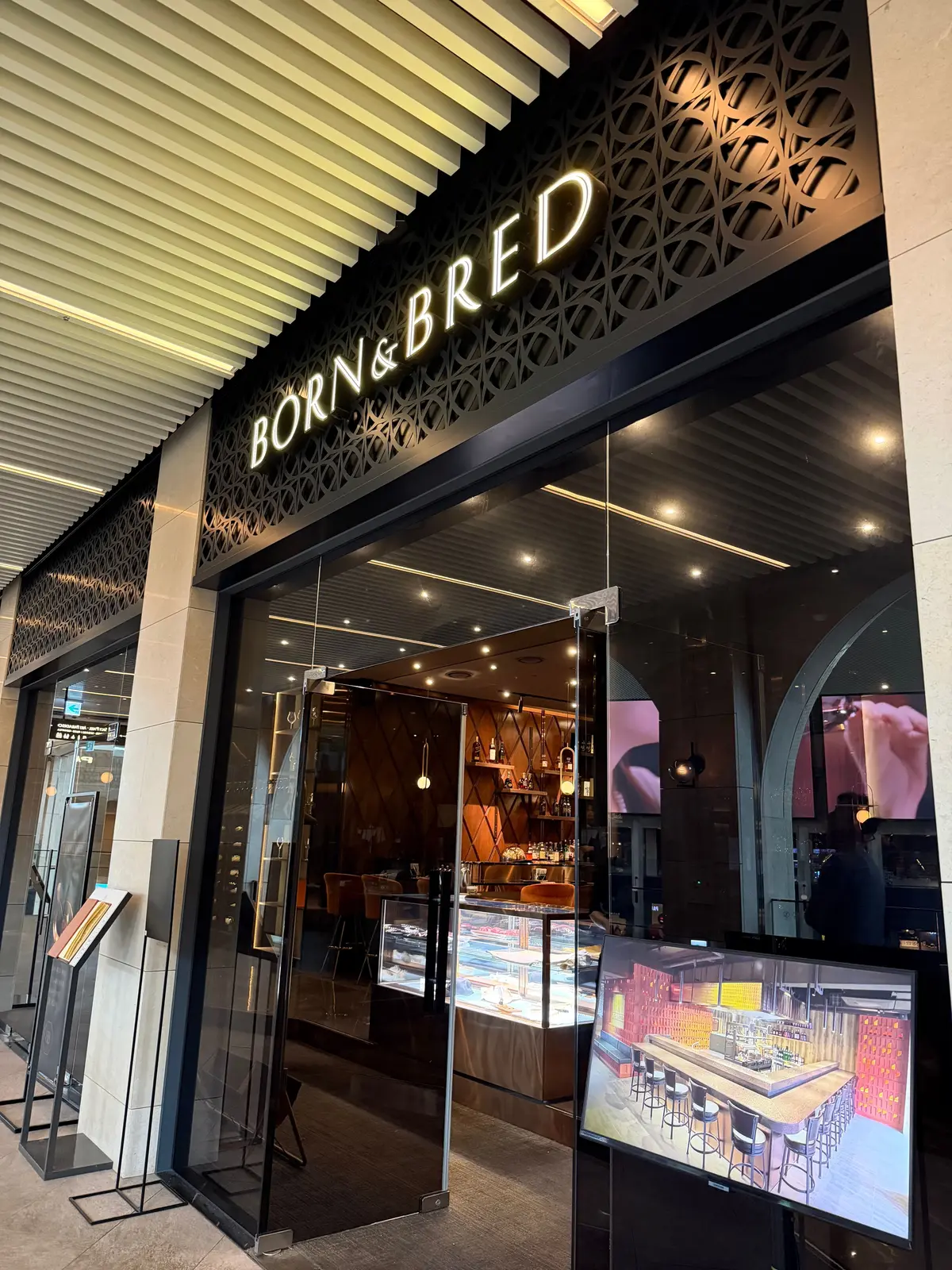 「BORN&BRED」の店頭。ガラス張りのエントランスから店内が見え、明るくモダンなインテリア、バーカウンター、椅子、ディスプレイケースが配置されている。外にはメニュー表が設置されている。
