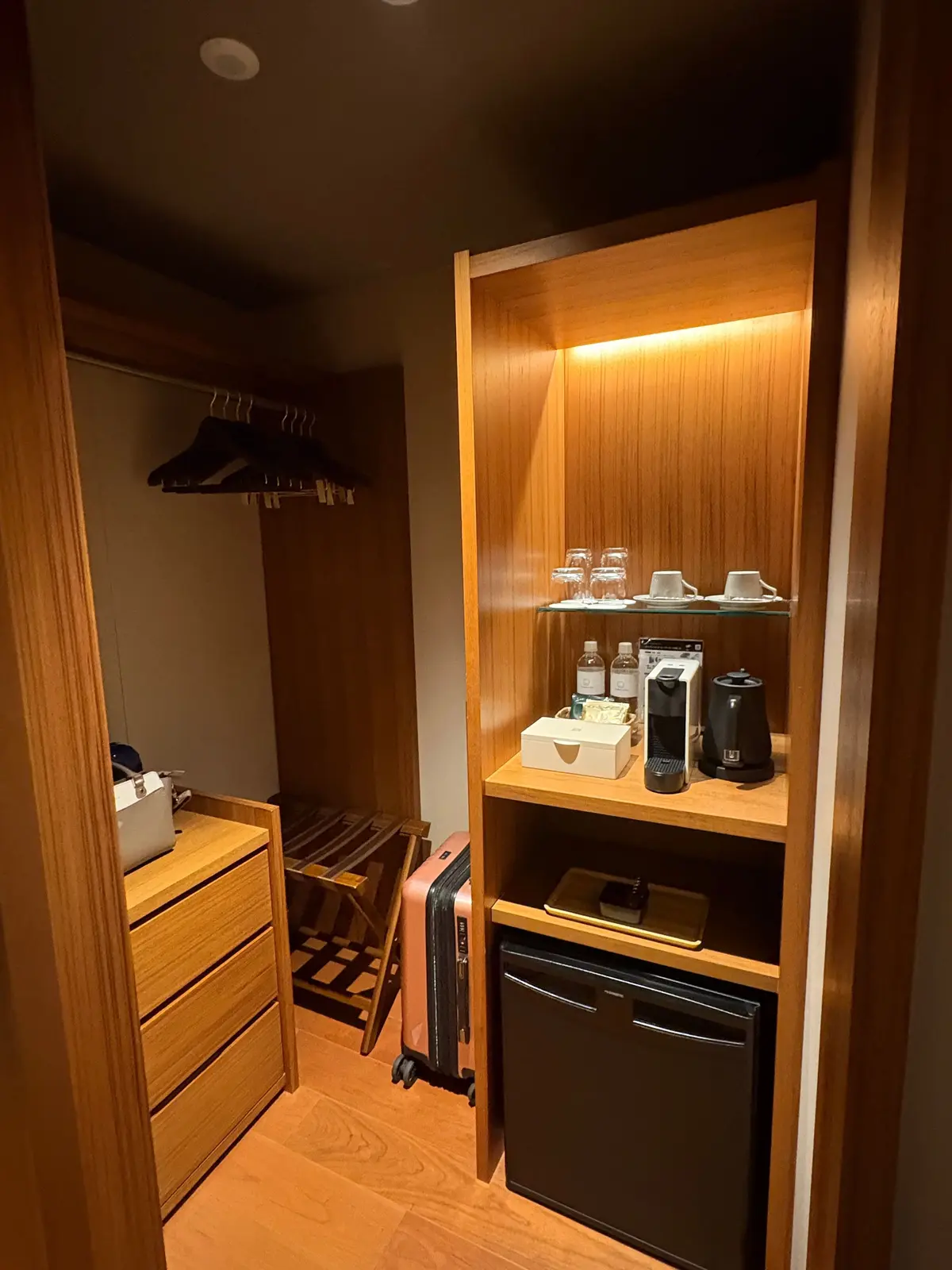 木目調の部屋のクローゼットとミニバー。左にはハンガーと引き出し、右にはネスプレッソ、グラス、冷蔵庫が設置されている。ピンク色のスーツケースがクローゼットの前に置かれている。