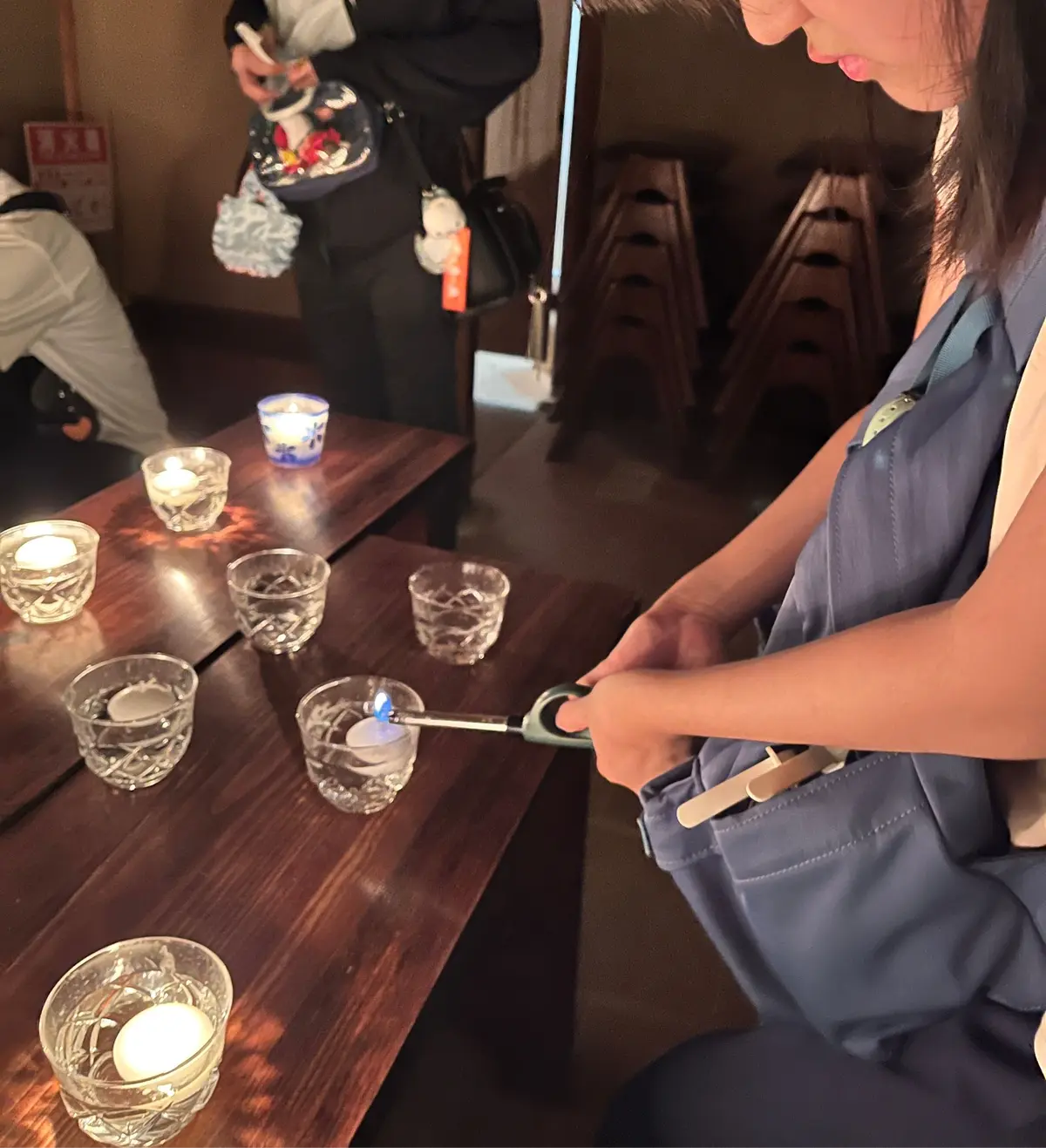 木製のテーブルに置かれた小さなガラスのキャンドル。女性がライターでキャンドルに火を灯している。