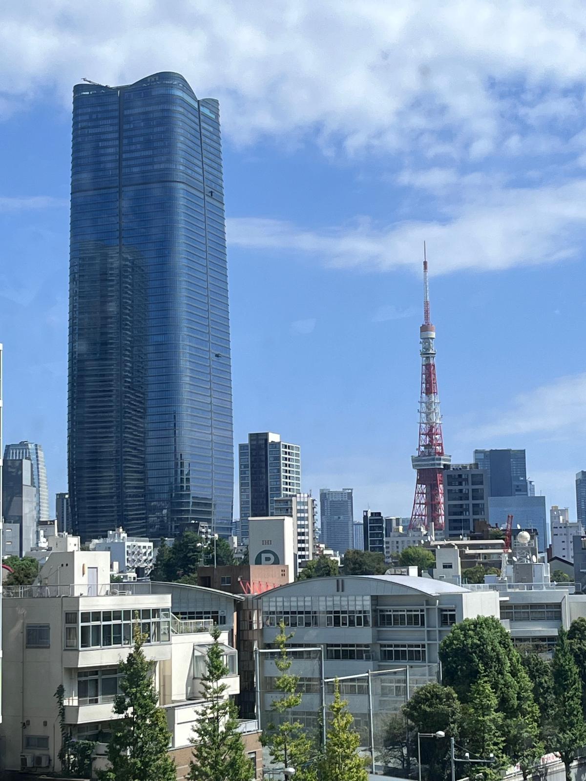 高層ビル群と東京タワーがそびえ立つ都市の風景。左には青いガラス張りの高層ビル、右には赤い東京タワーが目立つ。手前には緑豊かな木々や建物が配置されている。