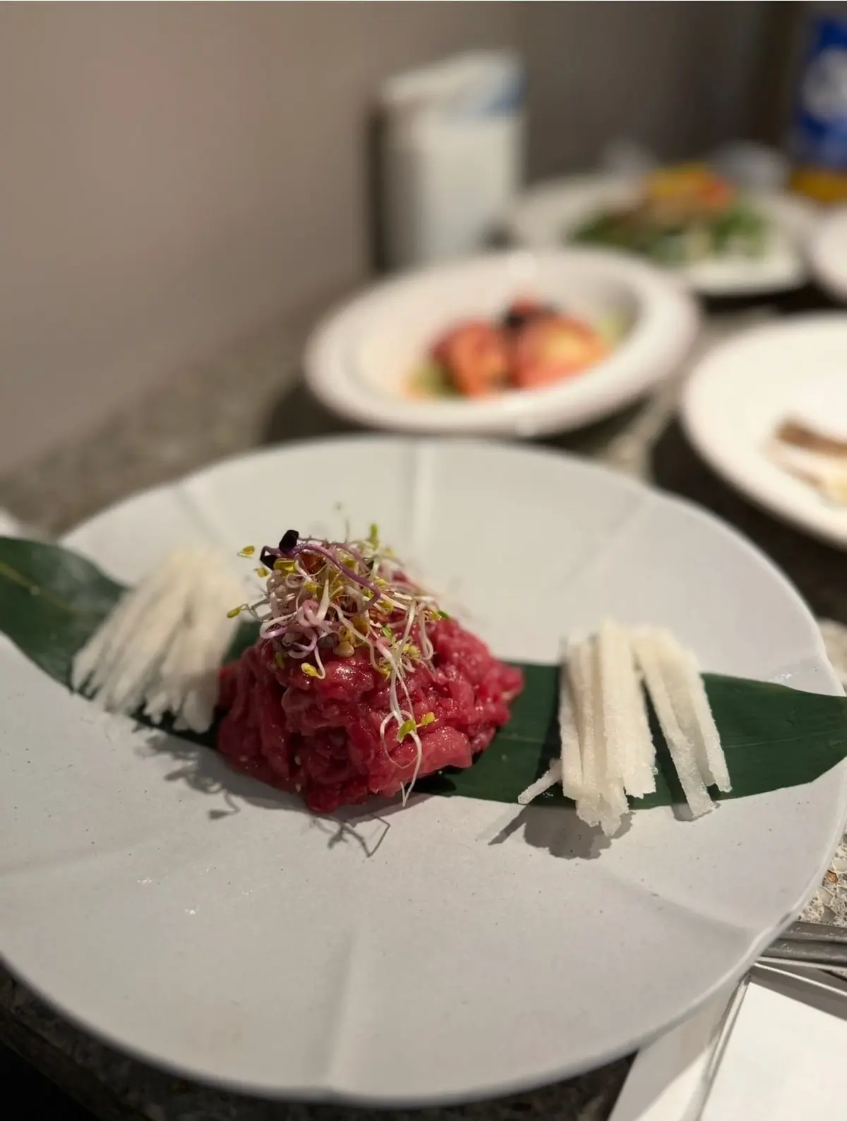 白いプレートに、緑の葉の上に赤身の肉のタタキが盛り付けられ、マイクログリーンの芽や白い細長い野菜が添えられている。