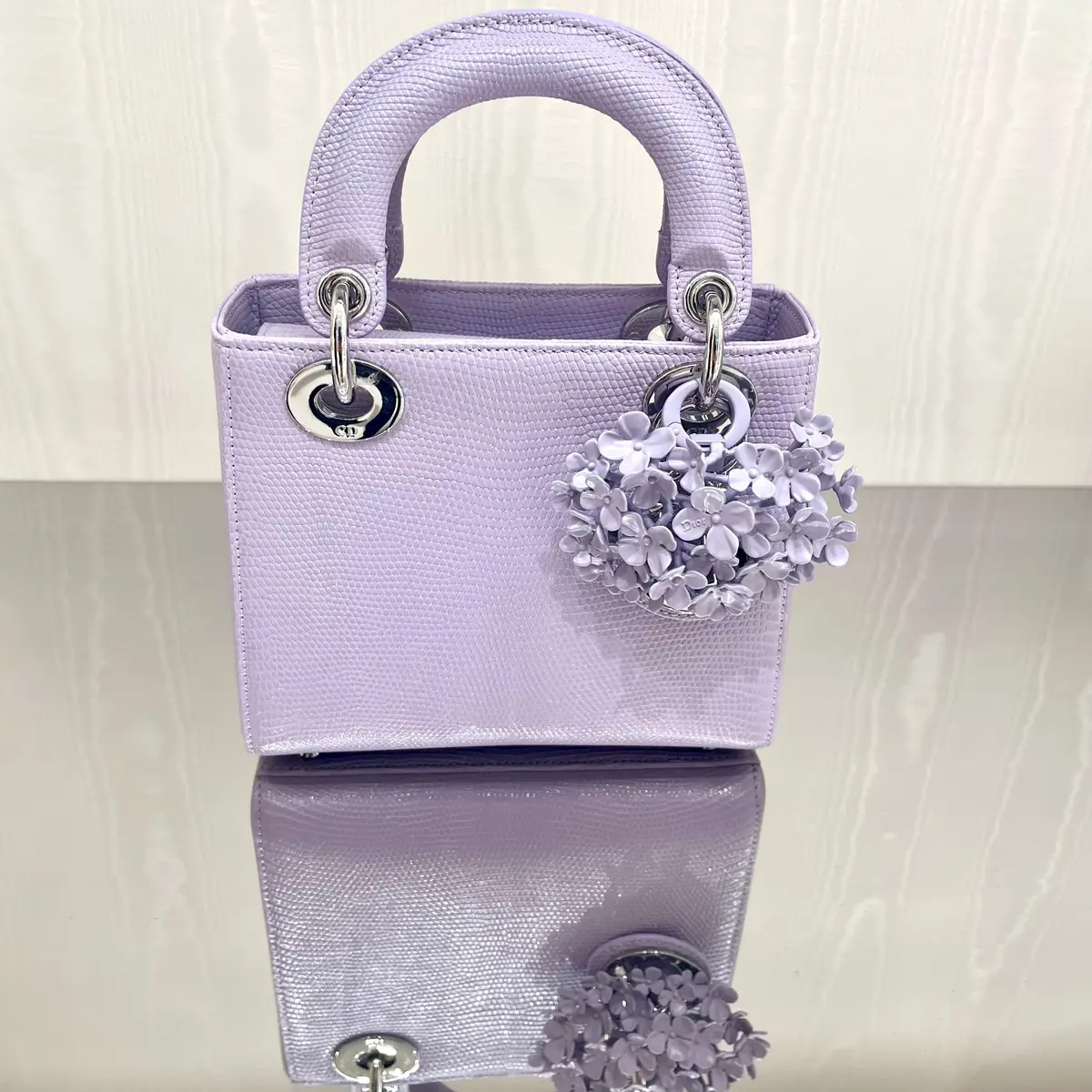 ディオールの淡い紫色のレディバッグ。シルバーの金具で装飾され、花柄のチャームが取り付けてある。光沢のある白いテーブルの上に置かれ、反射が見られる。