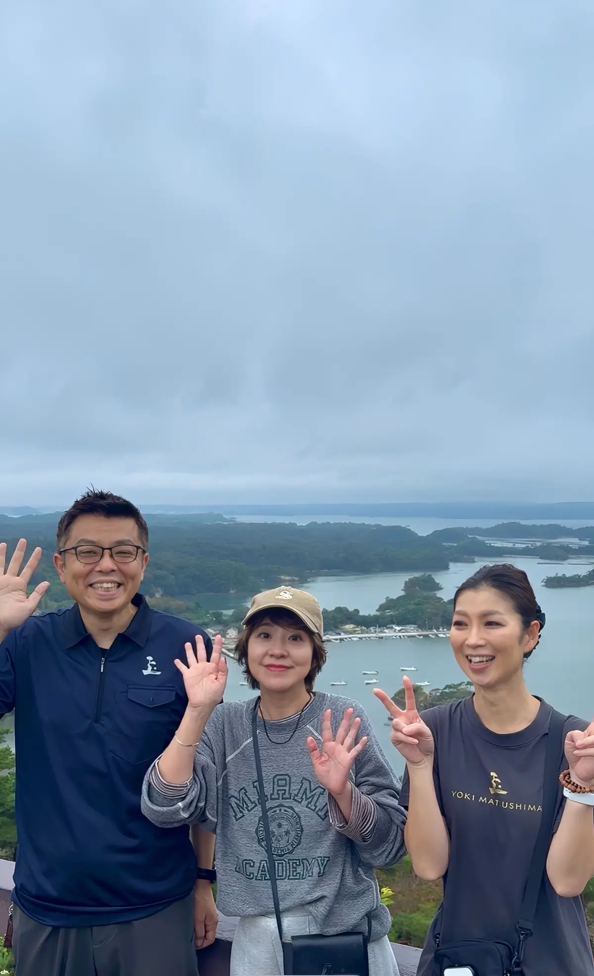 3人組が屋外の崖の上で、水面と緑豊かな島々を背景に手を振って笑顔を見せている。左から男性、女性、女性。空は曇り。
