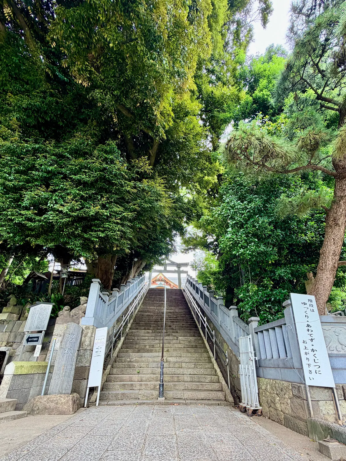 石造りの階段が、緑豊かな木々の間を縫うように上へと続いており、先には鳥居が見える。階段の脇には石碑や案内板が設置されている。
