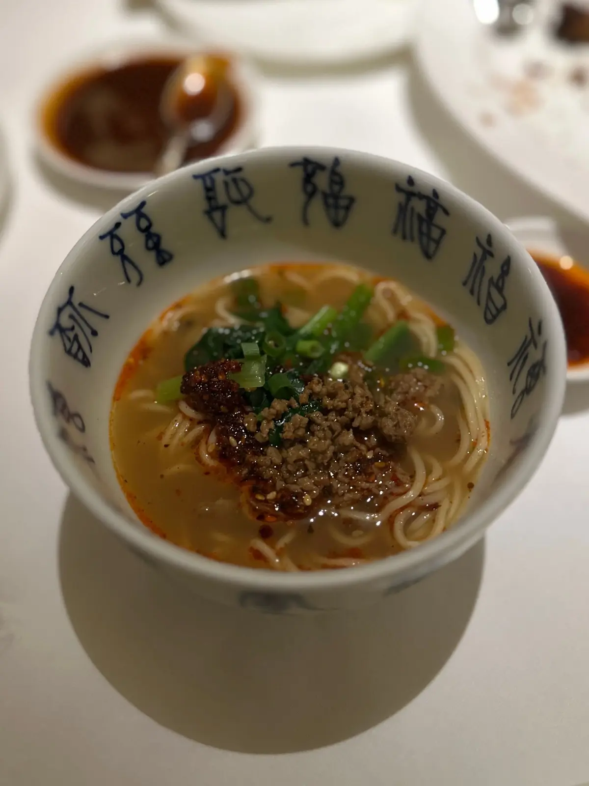 白いテーブルに置かれた、麺と具材の入った白い器。スープは淡い黄色で、ネギと黒い粒状のものが浮かんでいる。器の側面には青い文字が書かれている。