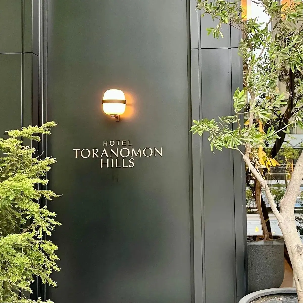 黒い壁に設置された「ホテル トラノモン ヒルズ」のサイン。上部には丸い壁掛けライトが付いており、左右には緑の植栽がある。