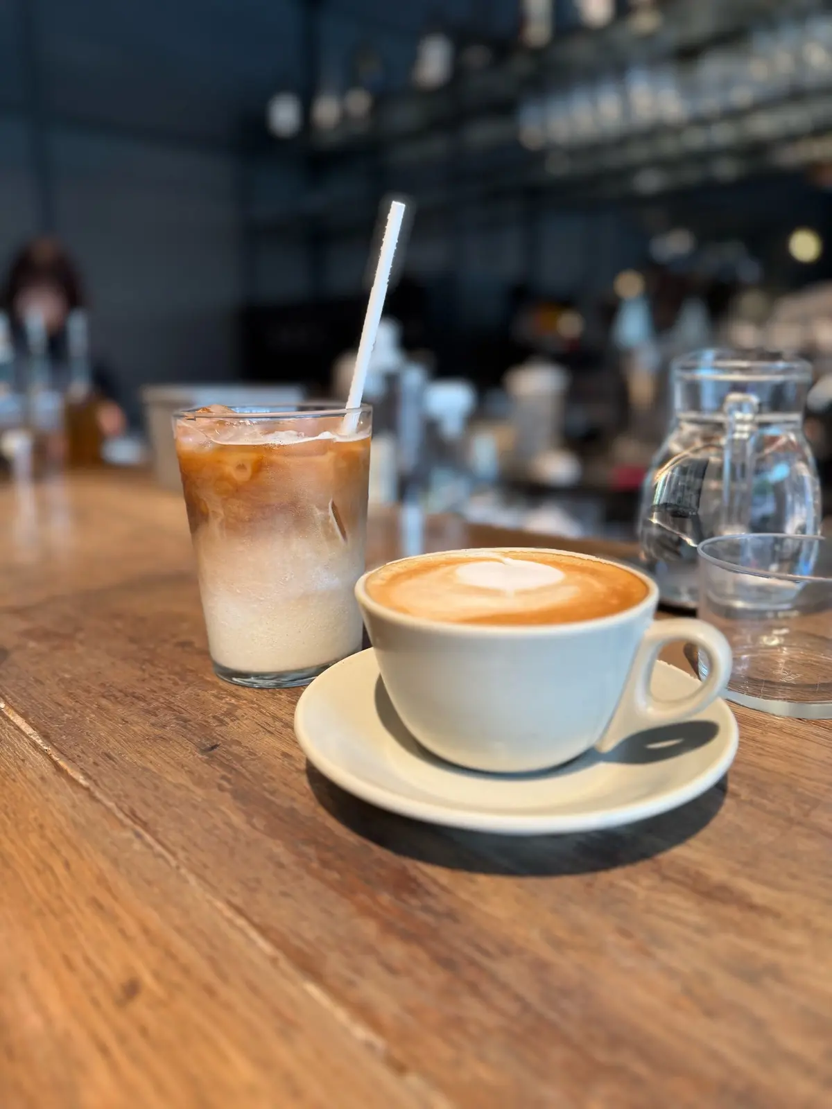 表参道カフェレストラン、RACINES AOYAMA、木製テーブルにアイスコーヒーとカプチーノが置かれている。アイスコーヒーはガラスカップに氷とストローが入っており、カプチーノは白いマグカップにハート模様のフォームが乗っている。