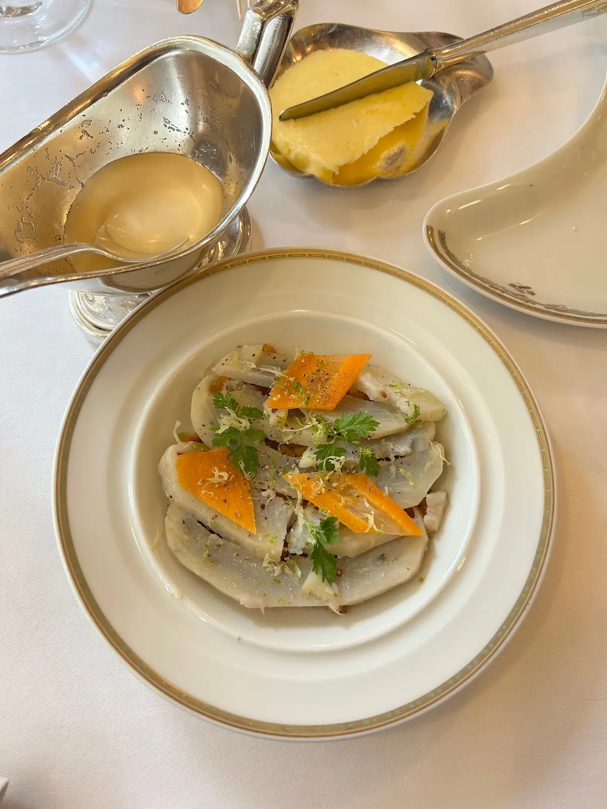白いプレートに、薄い色の魚の切り身とオレンジ色の野菜、緑のハーブが盛り付けられた料理。周囲にはスープとバターが置かれている。