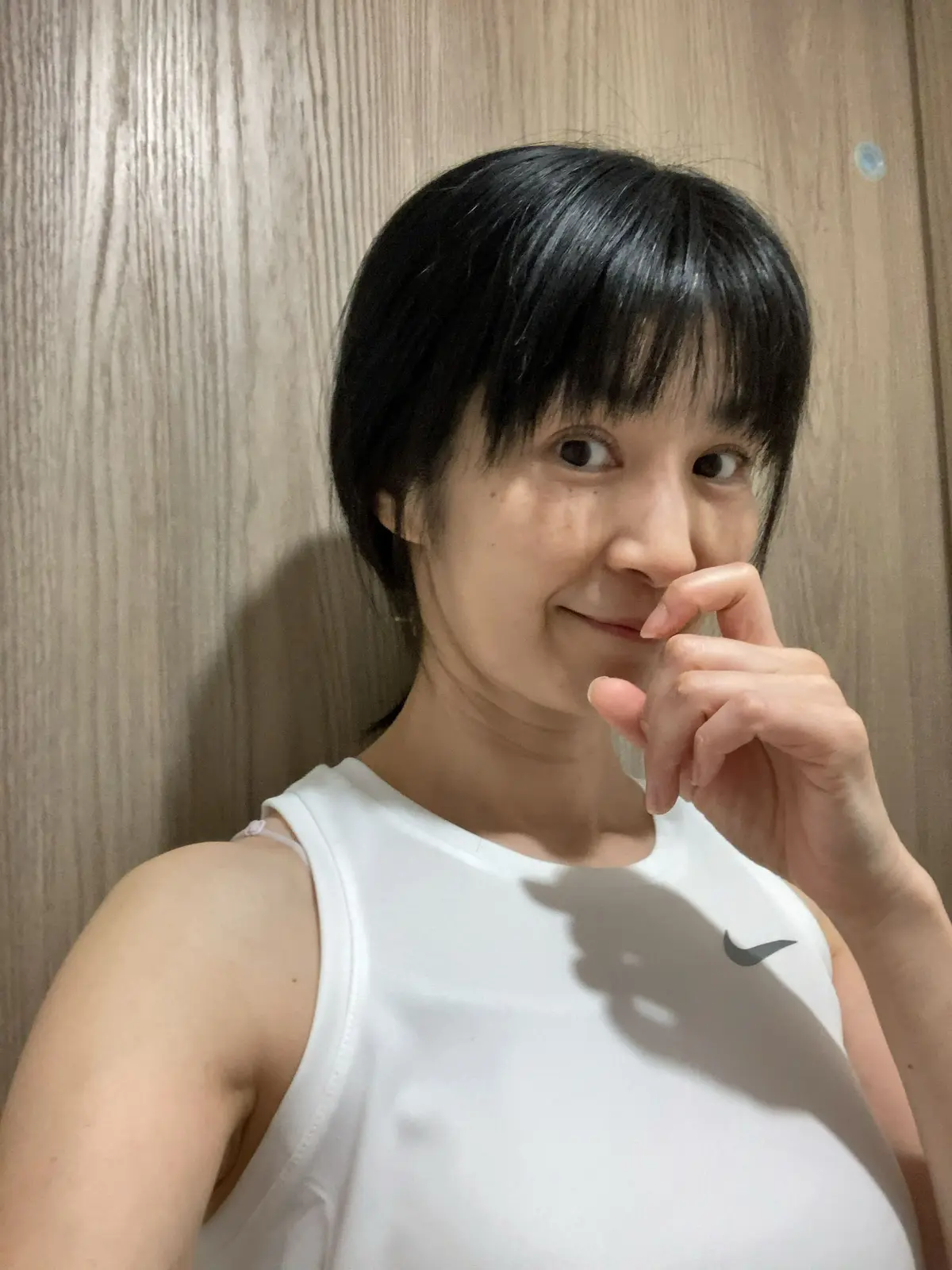 白いタンクトップを着た女性が、右手を頬にあてて微笑んでいる自撮り写真。黒髪のボブカットで、背景は木目調の壁。胸元にNikeのロゴが入っている。
