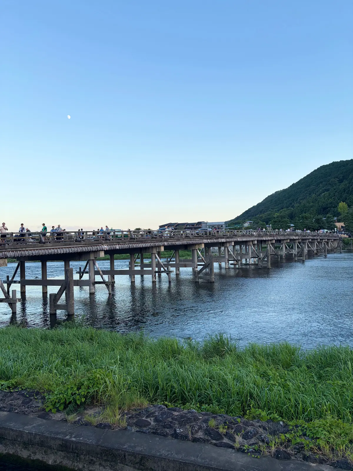 木造の橋が川に架かり、多くの人が橋の上でくつろいでいる。背景には緑の山がそびえ立ち、空には細い月が見える。手前には草が生い茂っている。