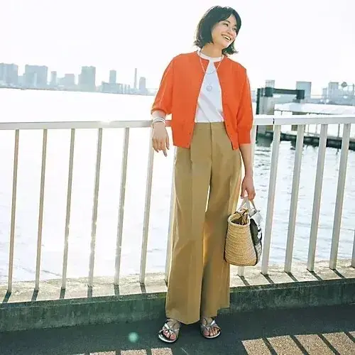 オレンジのオープンカーディガン、白いトップス、ベージュのワイドパンツを着た女性が、 waterfrontのフェンスに寄りかかり、ストローハンドバッグを持ち、笑顔で都市景観を眺めている