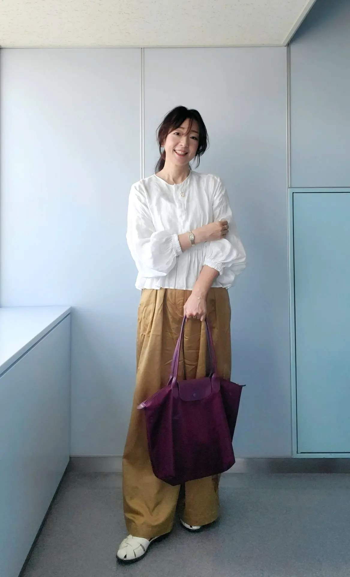 白いフリル袖ブラウスに、ブラウンのワイドパンツを合わせた女性。紫色のトートバッグを持ち、微笑んでいます。背景は淡いブルーの壁。