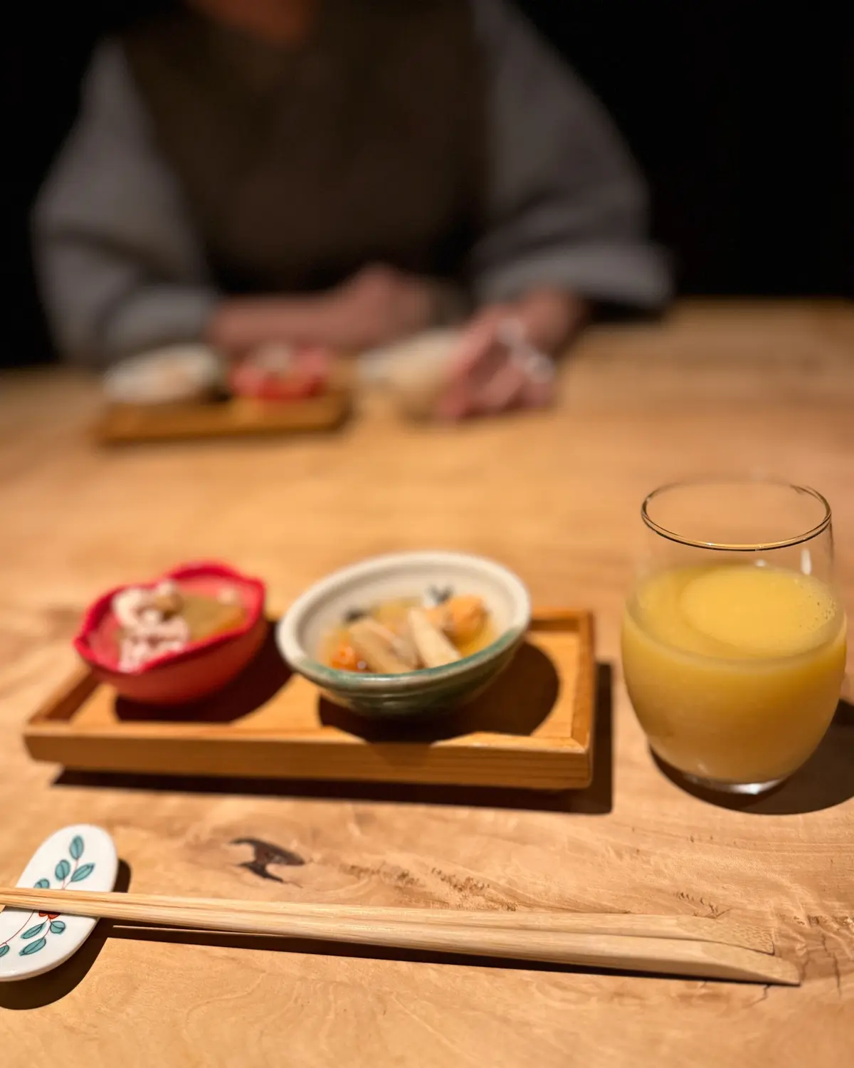 木製のトレイに、日本料理の盛り合わせと飲み物が置かれている様子。茶碗蒸し、焼き魚、お漬物、グラスに入った黄色い飲み物。背景には白い布が見える。