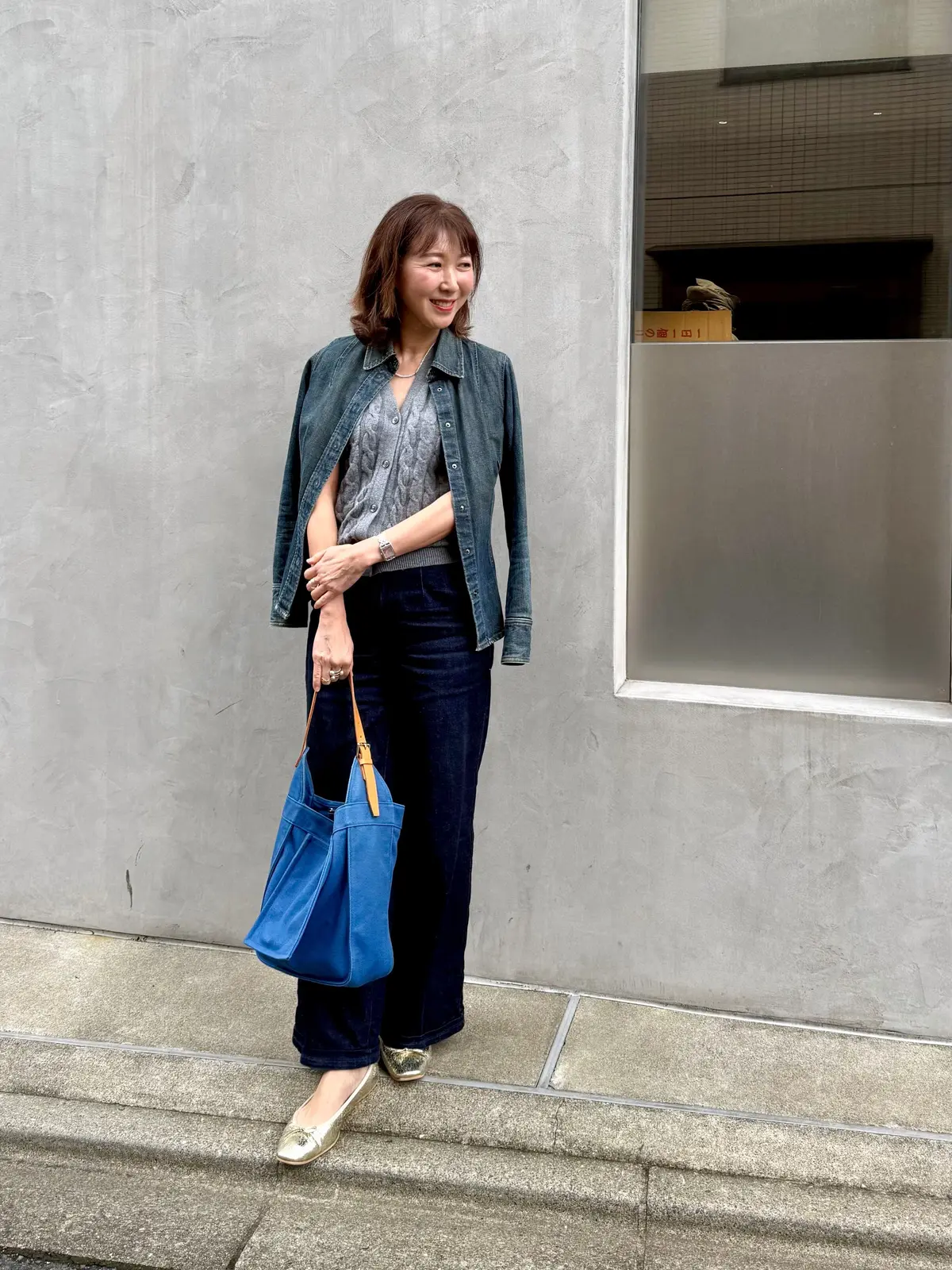 青いバッグを持つ女性。グレーの壁の前に立ち、微笑んでいる。ダークブルーのベルベットパンツ、グレーのデニムジャケット、白とシルバーのトップスを着用。足元はゴールドのパンプス。