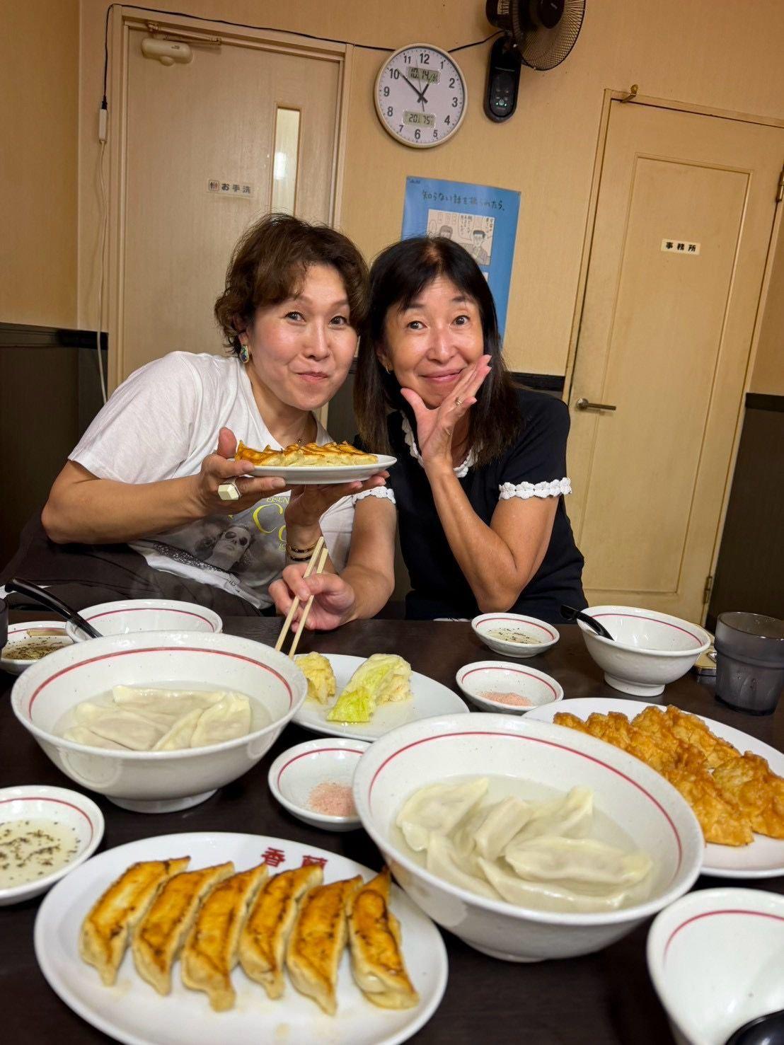 笑顔の女性2人が、たくさんの料理が並ぶ食卓を囲む。手前には餃子や唐揚げ、奥には麺類やサラダが白い皿に盛られている。壁には時計と案内板が掛かっている。