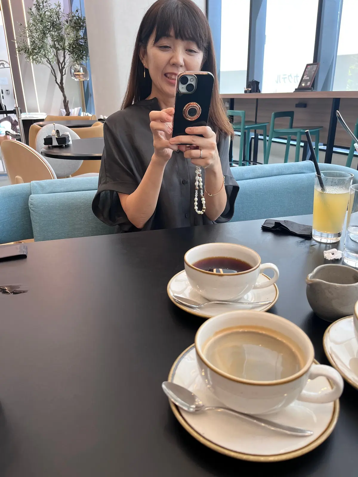 レストランで、女性がスマートフォンで自撮りをしている様子。黒いトップスを着ており、テーブルにはコーヒーのカップが置かれている。背景には窓から光が差し込み、モダンな内装が見える。