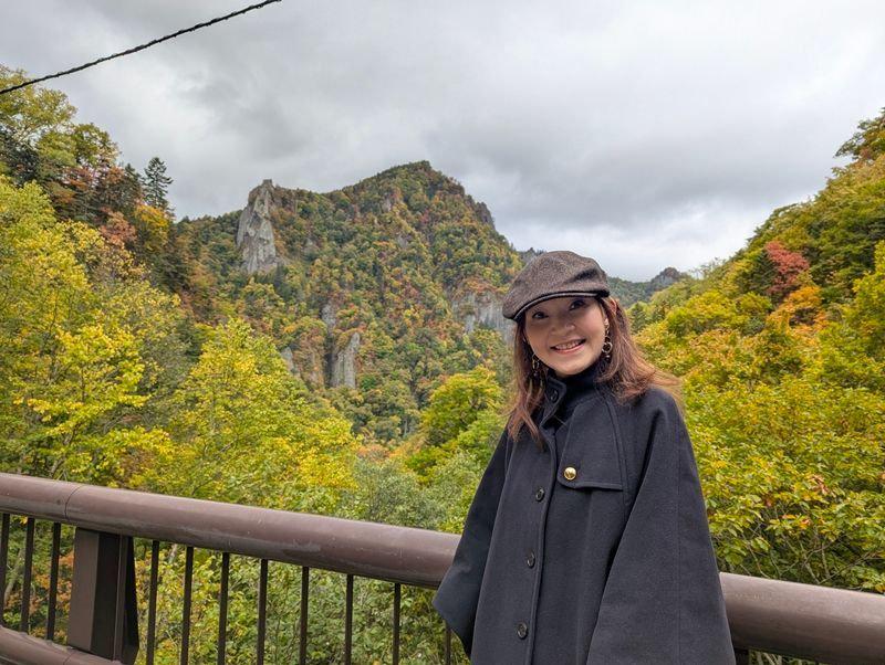 秋の山を背景に、黒いコートと帽子を身につけた女性が微笑む。手すりの前に立ち、右手にカメラを持っている。山の斜面は紅葉で彩られ、空は曇り空。
