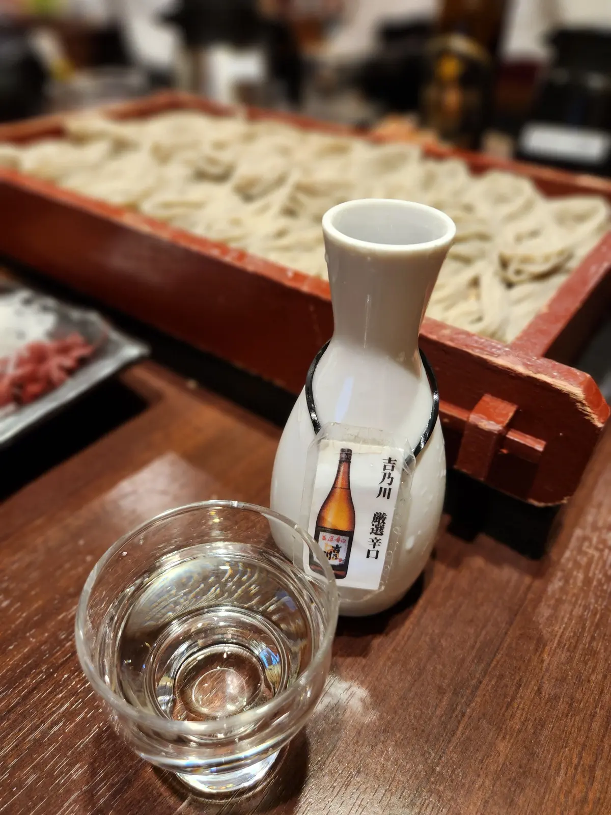 木製の盛皿に盛られたうどん、その横に白い徳利とグラスに入った日本酒。徳利には「吉力川 醸造辛口」のラベルが貼られている。