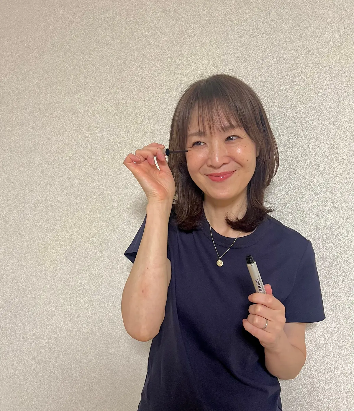 青いTシャツを着た女性が、ラッシュアディクトのまつげ美容液を持ち、片方の手で開けている。笑顔でこちらを見ている。