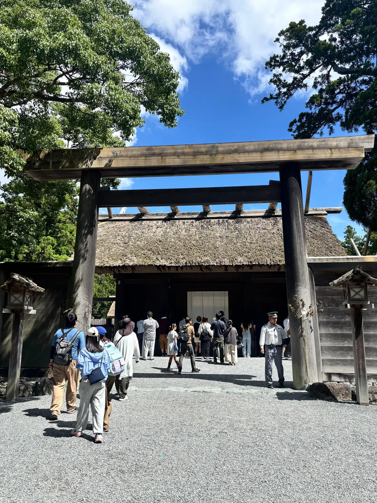 木造の鳥居と藁葺き屋根の建物。石畳の広場に人々が立っている。青空を背景に、緑豊かな木々が生い茂る。