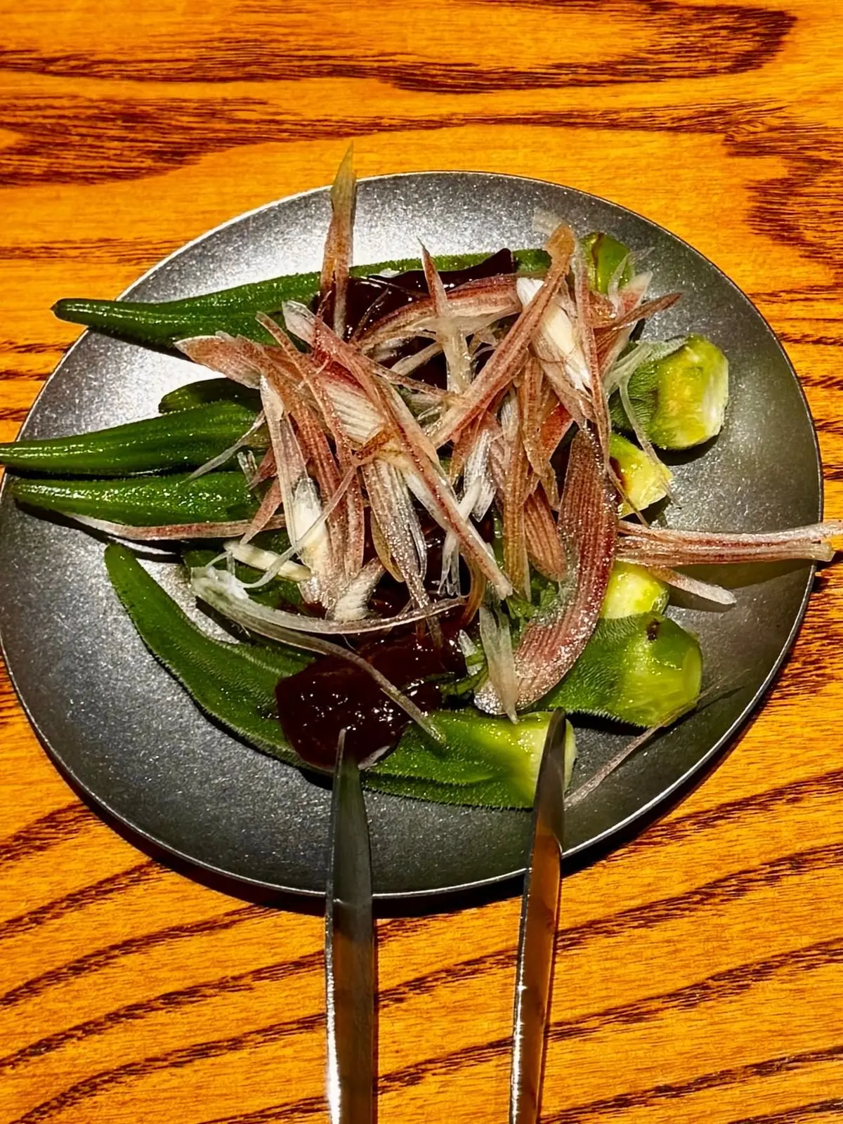 グレーの小皿に盛り付けられた、緑色の青梗菜と、薄切りになった赤色の根菜（おそらく人参）の和え物。中央に箸が置かれている。
