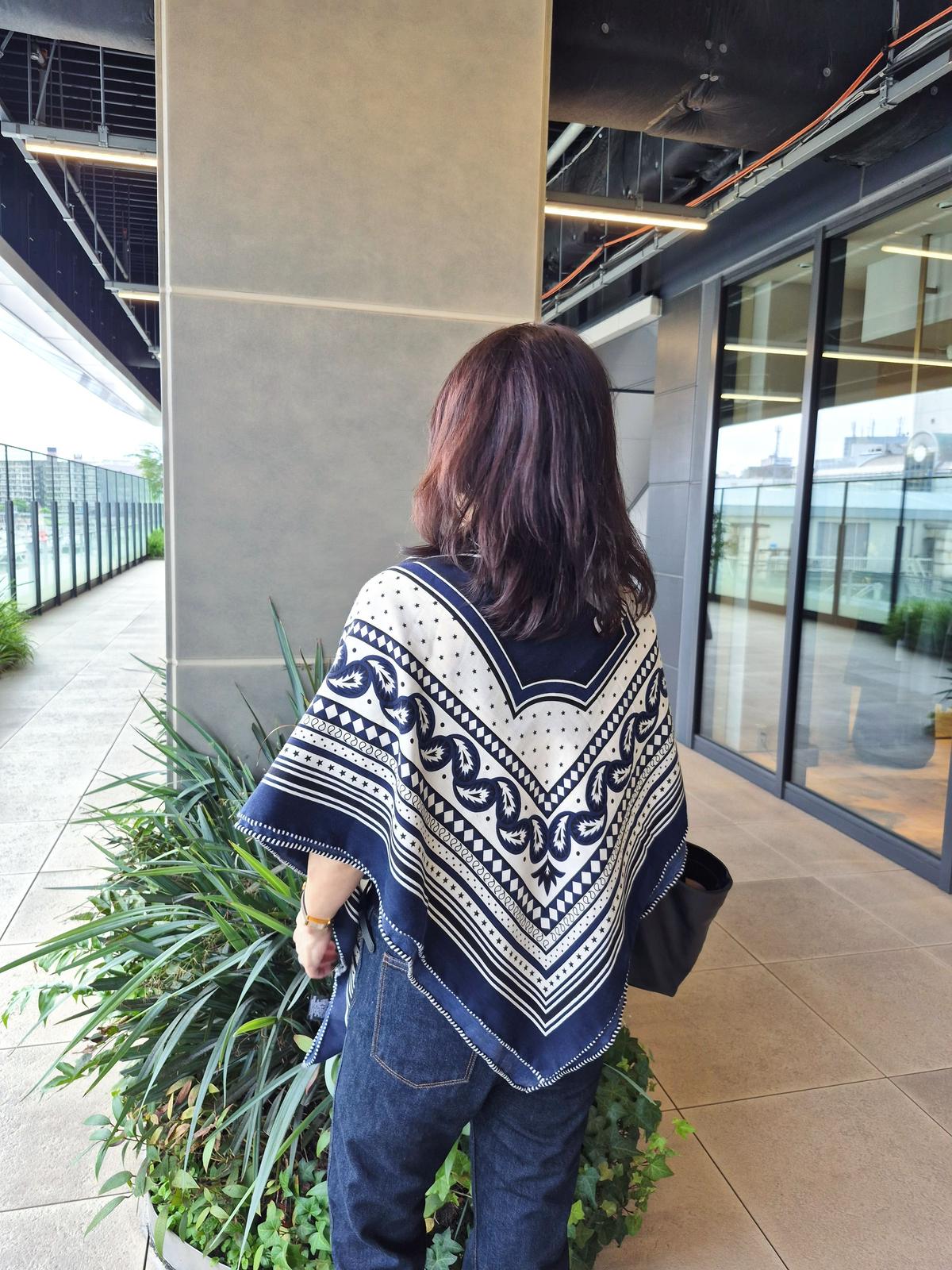 濃い青と白の幾何学模様のポンチョを羽織った女性が、タイル張りの廊下を歩いている。後ろ姿で、ダークブラウンの短髪。左手にはバッグを持ち、手前に緑の植栽がある。