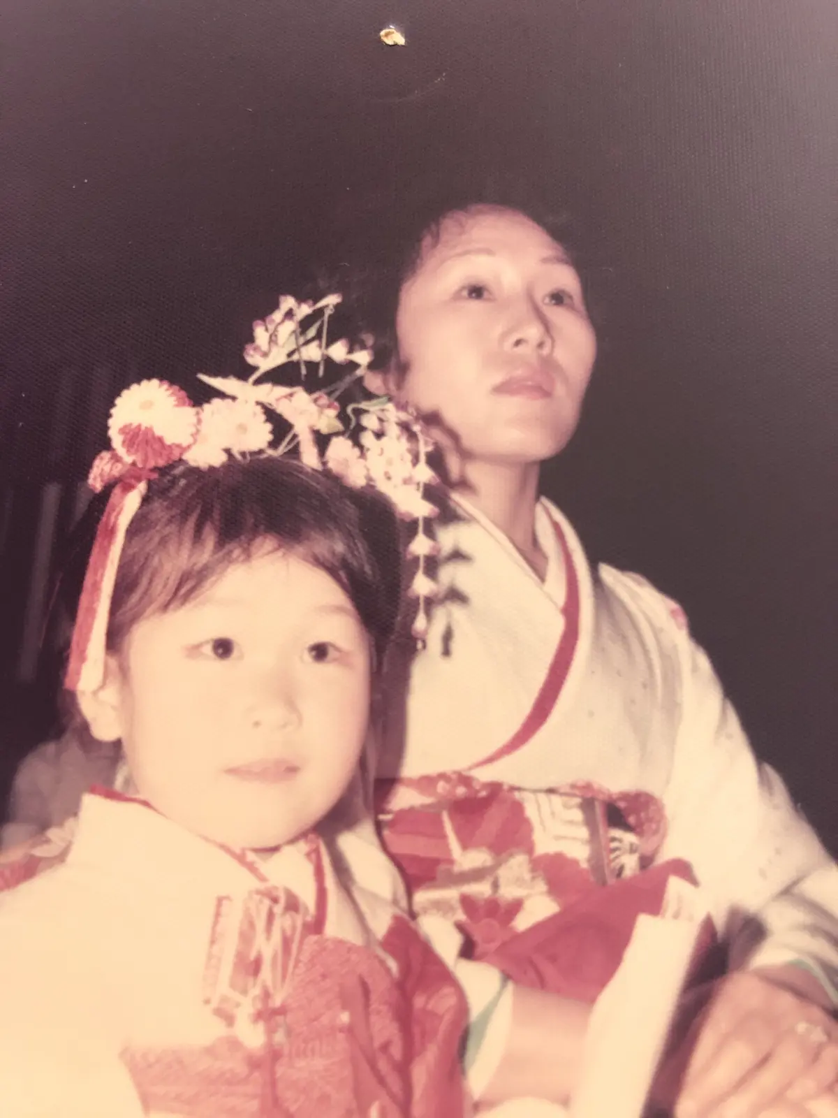 白地に赤縁の着物姿の母と娘。娘は花飾りをつけ、母は髪飾りを付けている。背景は暗く、人物が中央に配置されている。