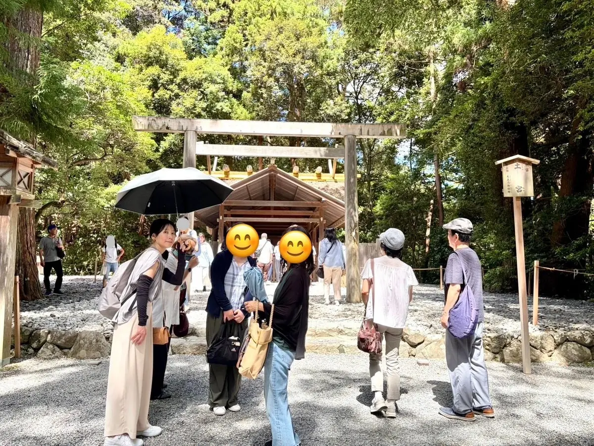 木々に囲まれた鳥居の前で、複数の人が会話している様子。女性が傘を持ち、他の人はカジュアルな服装。背景には石畳の道と緑豊かな木々が見える。