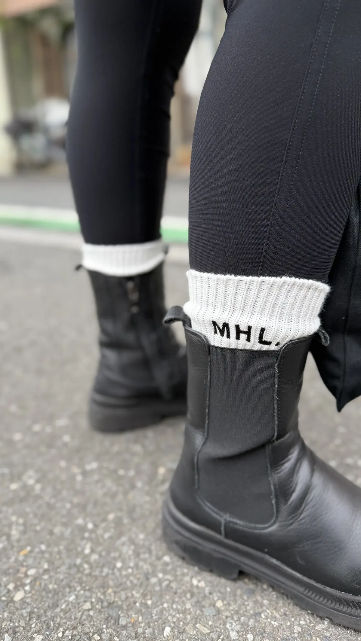 黒いレギンスと黒いチェルシーブーツを履いた足元。白い靴下で「MHL.」のロゴが刺繍されている。背景には道路と緑色のラインが見える。