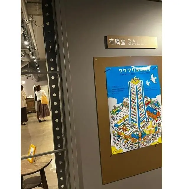 灰色の壁に「有隣堂GALLERY」のサインと、青と黄色のイラストポスターが飾られている。ポスターには高層ビルと人々が描かれ、奥には展示会場への入り口と来場者がいる。