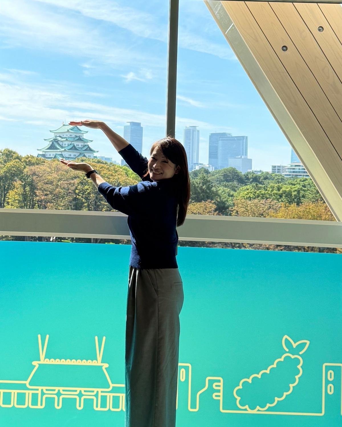 青い壁の前で、女性が両手を広げ笑顔。背景には、青空を背景にした城と高層ビル群が見える。壁には白い線で描かれた日本のシンボルが描かれている。