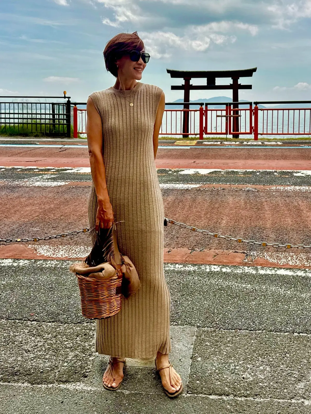 茶色のリブニットのロングドレスを着た女性が、鳥居を背景に立っている。サンダルを履き、編みバッグを持ち、サングラスをかけている。背景には海と空が広がり、少し曇り空。
