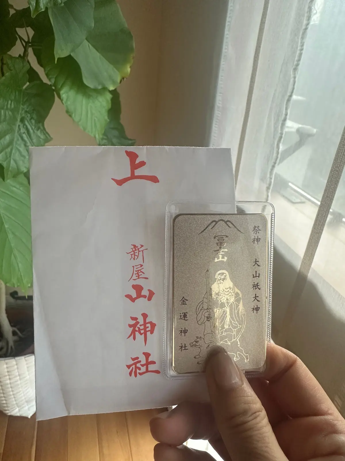 白い紙に赤い文字で「上」「新屋山神社」と記載されたお守りの紙と、プラスチックケースに入った金属製の神像のお守りが手の中に置かれている写真。背景には植物と窓が見える。