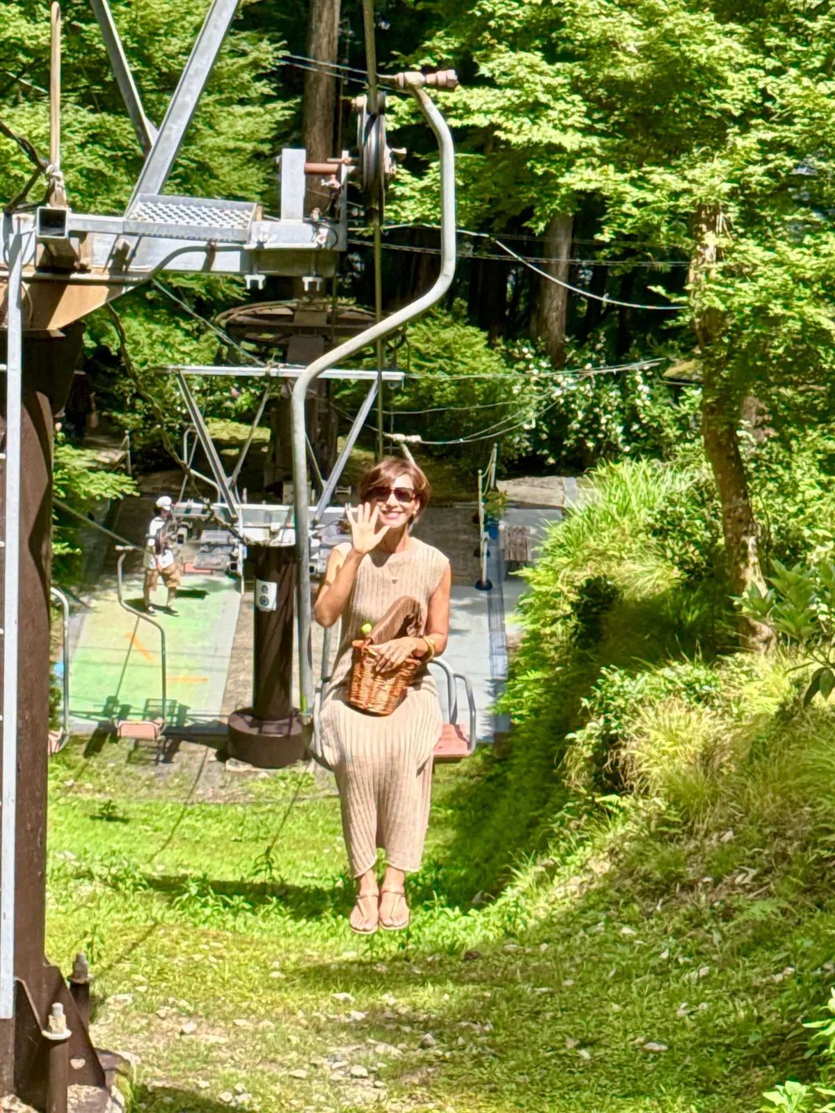 緑豊かな木々の背景に、ベージュのワンピースを着た女性が笑顔で手を振っている。彼女はサングラスをかけ、籐のバッグを持っている。背後には、スキーリフトの支柱とケーブルが見える。
