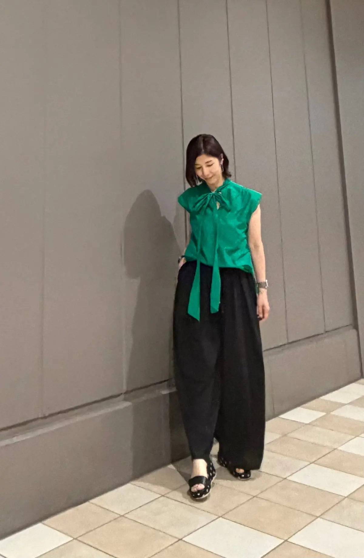 緑色のノースリーブブラウスと黒いワイドパンツを着用した女性が、グレーの壁の前に立っている全身写真。ブラウスは襟元にリボン結びがあり、黒いサンダルを履いている。