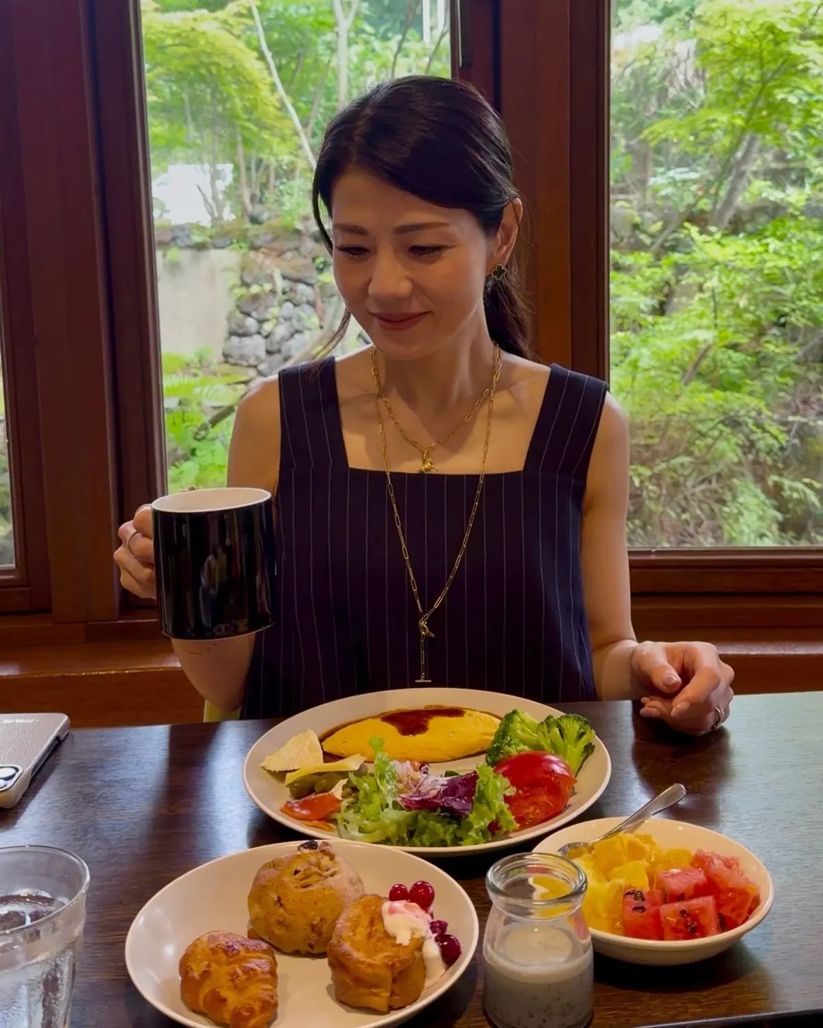 窓際に座る女性が、サラダとオムレツ、焼き菓子、フルーツの朝食を楽しんでいる様子。黒いマグカップを持ち、金色のネックレスを着用。背景には緑豊かな自然が見える。