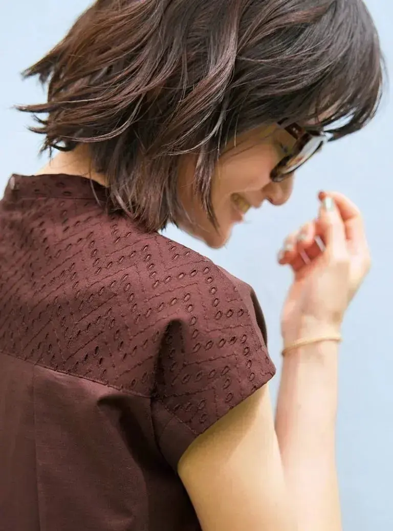 茶色のトップスを着た女性が、こちらを振り返っている。茶色の短髪で、眼鏡をかけている。右腕を前に出し、指先で何かを確かめている。背景は青色。