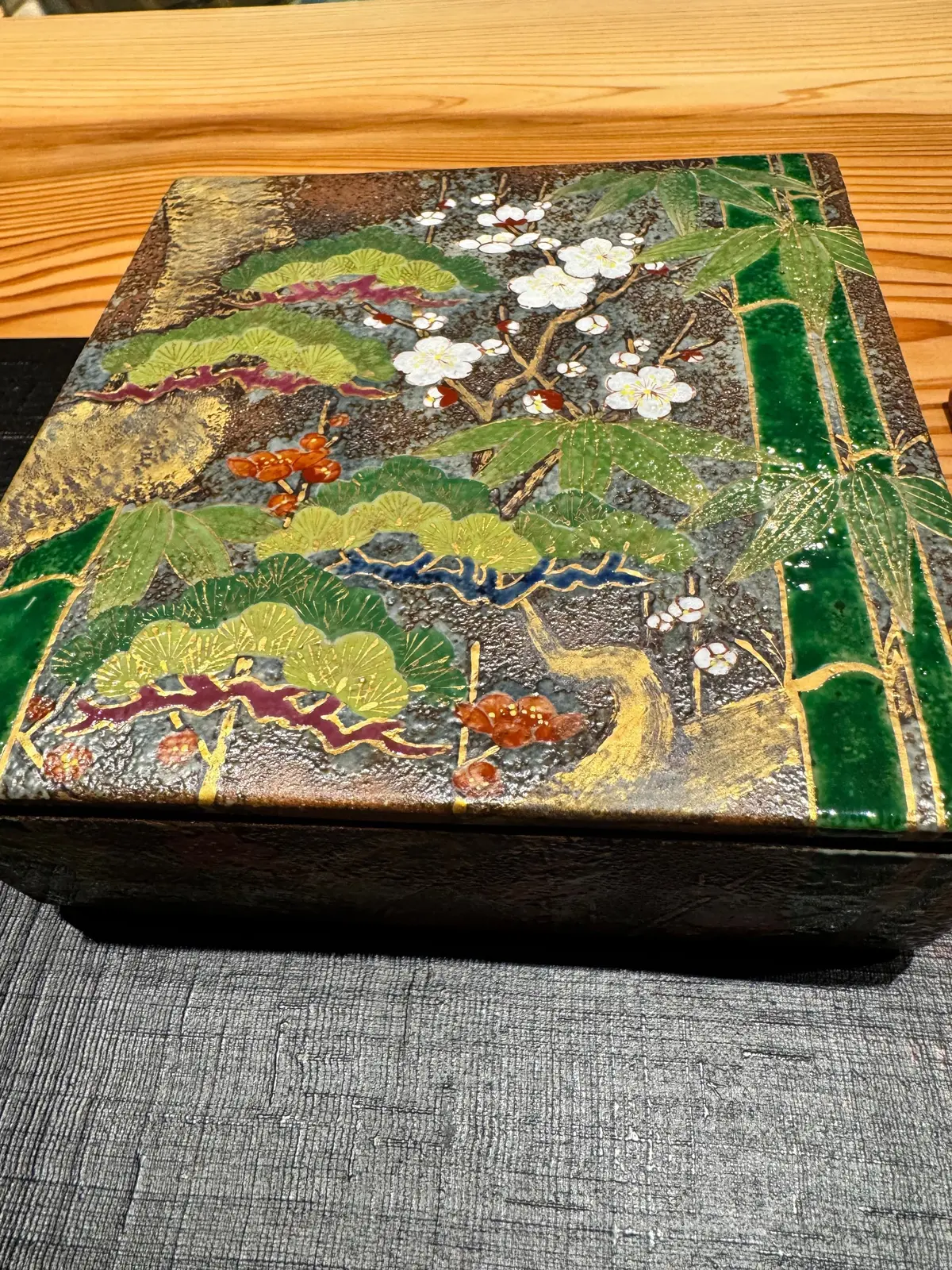 漆塗り箱の蓋に、白花、緑の葉、竹、赤褐色の実などが描かれた風景模様。金彩が施され、黒い背景に鮮やかな色彩が映える。