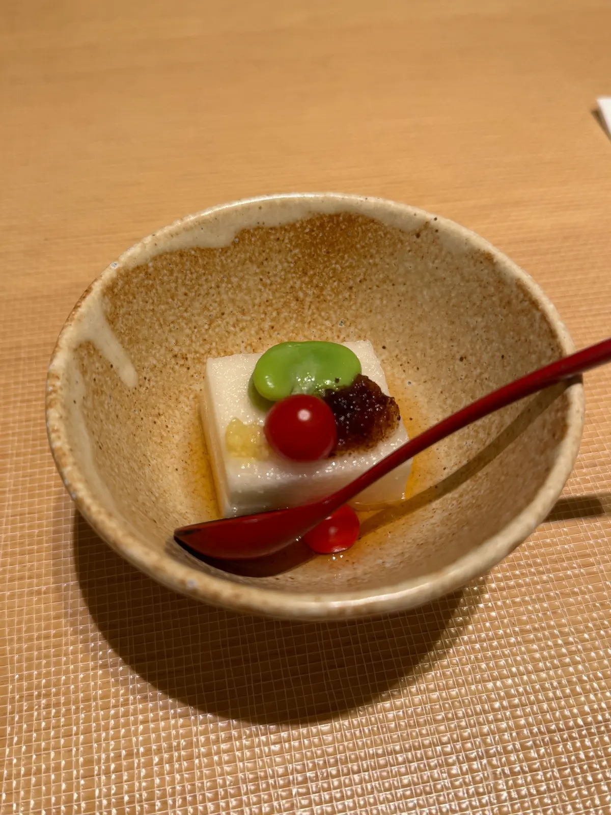 茶色い土器の器に、豆腐の上に緑の豆、赤いチェリー、黒いソースがのせられた和菓子。赤いスプーンが横たわる。